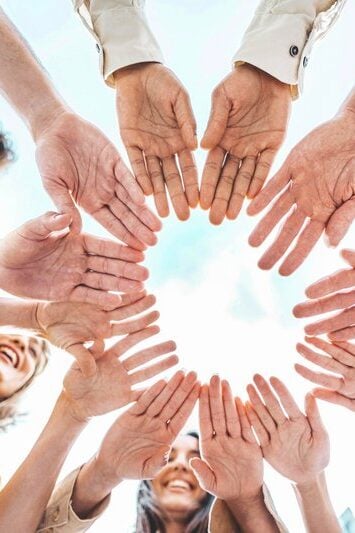 A group of young, diverse individuals forming a circle with their hands outdoors, symbolizing unity and community support.